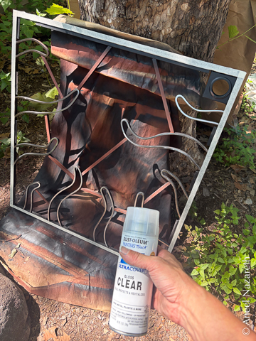 The tentacled frame is leaning against the tree. The edges are masked with masking tape. A can of clearcoat spray is held in front of it.